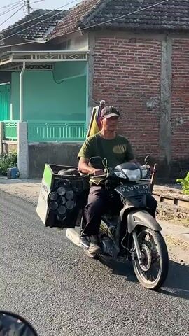 A man rides a black motorcycle on a paved road. He wears a dark green t-shirt, dark pants, and a dark baseball cap. He is carrying a large container attached to the back of the motorcycle. The container is black with a white label and has six circular openings on its side, each containing a cylindrical object. The man appears to be looking forward. The environment is a street lined with houses, one of which has green siding and a white fence, and another with a brick facade. The sun is shining, casting shadows.