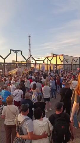 A large crowd of people are gathered outdoors, likely at an event or festival. They are facing a raised platform or stage area where performers are visible, though not clearly discernible. The crowd is dense and spans across the frame. The setting appears to be urban or industrial, with buildings and a tall antenna or tower in the background. The sky is a clear blue with some wispy clouds, suggesting it is daytime. The lighting indicates it could be late afternoon or early evening. People are dressed casually in a variety of clothing, and some are holding up phones, possibly to record the event. The overall atmosphere appears to be one of engagement and enjoyment.