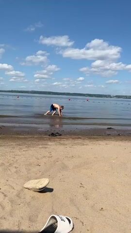 A boy wearing blue swim trunks and goggles is in shallow water at the edge of a sandy beach. He is lying on his stomach, moving forward into the water and appears to be swimming. The water is calm and clear. In the background, there is a body of water with buoys marking a swimming area. The sky is blue with scattered white clouds. There are a pair of black flip-flops and a single white shoe on the sand.