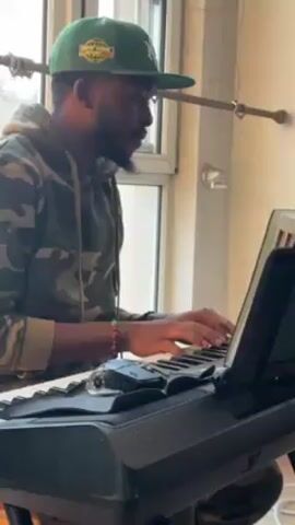 A man wearing a green baseball cap and camouflage hoodie plays a digital keyboard in a bright room. The keyboard, which has a sheet music stand, sits on a black metal stand. Next to it on a wooden chair is a laptop. A microphone stands near the keyboard. Large windows let in natural light, illuminating the room and its hardwood floor.