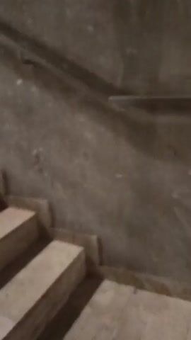 The camera is looking up a set of beige-colored stairs. The stairs appear to be made of marble or a similar material. A metal handrail is visible on the right side of the stairs, and the wall is a similar beige color with a rough texture. The lighting is dim, and the upper portion of the staircase and the landing above are dark.  The camera is shaky, suggesting it is being held by someone moving up the stairs.