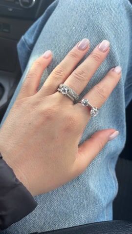 The video shows a woman's left hand resting on blue denim jeans. The hand wears two rings. The ring on the ring finger is a set of three silver rings, with the main ring featuring a center stone. The second ring is on the middle finger, a silver ring with two stones at each end. The woman moves her hand slightly. Background music is audible.