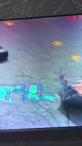 The video shows a robot combat competition. One robot, identified as MINOTAUR, is visible on the left side of the arena. Another robot, partially obscured, is on the right. The robots are engaged in combat, with sparks flying and the robots moving rapidly. The arena floor is marked with lines and has various obstacles or power-ups. The background appears to be a stadium or venue with audience seating, though no audience members are clearly visible. The lighting is dramatic, with colorful spotlights illuminating the combatants.