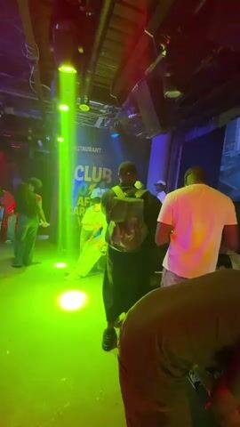 The video shows the interior of a nightclub. People are standing and sitting at tables, illuminated by multicolored lights, primarily green and red. A screen displays moving images of models or dancers, possibly as part of a music video or performance. The club has the words "CLUB OF AFRICAN CARIBBEAN" written on the wall. Patrons are dressed in various casual outfits. The camera moves around the space, capturing different angles of the people and the environment.