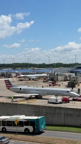 The video shows an airport tarmac on a sunny day with a few airplanes visible. The main focus is a silver American Eagle jet parked at a gate, with a jet bridge connected. Two people in yellow vests are visible near the front of the plane, and another person is near the tail. There are various service vehicles and equipment around the plane, including a white generator and a red cart. In the background, another American Eagle plane is parked at a different gate, and several other aircraft are visible further in the distance. The sky is blue with some scattered clouds, and trees and airport buildings line the horizon. A concrete barrier runs across the bottom of the frame, with a road and a bus visible in the foreground.