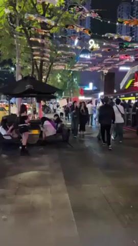 The video shows a public space filled with people at night. Overhead lighting provides illumination, highlighting the brick paving of the walkway. Many people are visible, engaged in various activities such as standing, walking, and sitting on benches. Two women in the foreground walk towards the camera while taking a selfie. String lights hang from trees above, and building facades feature signage like “9th Street”. The ambient noise suggests a lively environment with possible music or chatter.