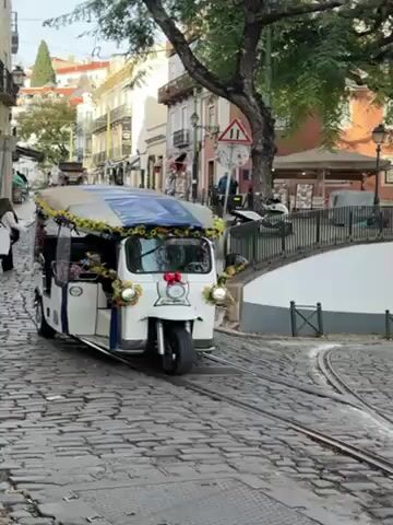 The video shows a street scene in Lisbon, Portugal. The street is paved with cobblestones and includes a tram track that forks into two directions. In the initial frame, a white tuk-tuk decorated with flowers, featuring a blue canopy and the word "FADO," is positioned on the right side of the frame. A woman is seated in the driver's seat. A person in a white coat crosses the street on foot. As the video progresses, the tuk-tuk moves out of view, and more pedestrians are seen walking across the tram tracks. A street sign, buildings, and a small park area are visible in the background. The sky is bright, suggesting a sunny day.