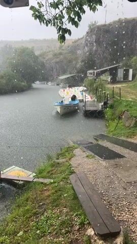 This video depicts a waterfront scene during heavy rainfall. The primary subjects are several small boats docked on a calm body of water, likely a river or lake, surrounded by lush green vegetation and a rocky cliff face in the background. Some of the boats appear to be paddle boats, identifiable by their colorful seats. A wooden dock is visible, with a few weathered wooden planks laid out on the shore in the foreground. Behind the boats, there's a building with a covered awning and a camper trailer parked nearby.

The activity taking place is simply the presence of these boats on the water under adverse weather conditions. The heavy rain is a very notable detail, with visible raindrops impacting the water's surface and creating a grey, hazy atmosphere. The scene suggests a location that is likely a recreational area, possibly a rental spot for boats, which is currently deserted due to the rain. The weather implies a gloomy or overcast time of day, with no direct sunlight visible. The rocky cliff and dense greenery point towards a natural, possibly somewhat remote, setting. The overall emotion evoked is one of tranquility interrupted by the elements, a sense of nature's power. No text is clearly legible in the video.