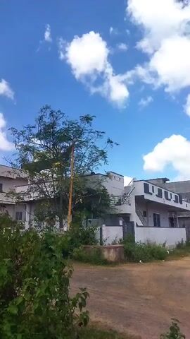 The video shows a suburban area with houses and construction sites under a blue sky with white clouds. In the foreground, there are bushes and dirt piles. In the background, there are houses, a tree, and electricity towers. The camera pans to the right, revealing more of the landscape and construction activity.