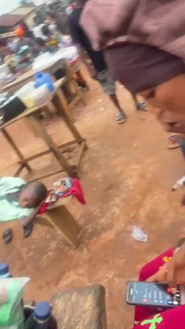 A young boy lies on a wooden bench, his head resting on his arm, in what appears to be a marketplace. He is wearing a green and white striped t-shirt and dark shorts. Several bottles and packaged goods are visible on a nearby table and in the foreground. In the background, other people are present, some on motorcycles, and stalls are set up. The ground is covered in reddish-brown dirt. The scene is outdoors, with overcast skies.