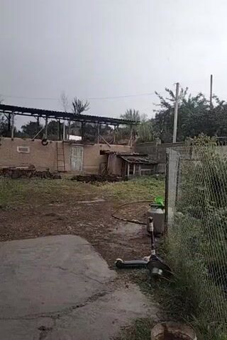 The video captures an outdoor scene under an overcast sky, suggesting potentially inclement weather. In the foreground, a black kick scooter lies on a concrete surface. Behind the scooter, a chain-link fence runs horizontally, bordering an area with dense green foliage. To the left of the fence, a large, grey cylindrical object, possibly a water tank, stands upright, with a green watering can perched on top. Further into the background, several structures are visible. One appears to be a weathered, single-story building with a light-colored door and a ladder leaning against its wall. Adjacent to it is a smaller, dilapidated structure with a sloped roof. A covered, open-sided structure with a corrugated roof is also present. Trees with dark green foliage are scattered throughout the scene, with a more substantial cluster of trees and bushes behind the fence. Utility poles are also visible in the distance. The ground appears damp and somewhat muddy in places, further indicating the possibility of rain. The overall impression is that of a rural or semi-rural backyard or compound experiencing a dull, possibly wet day.