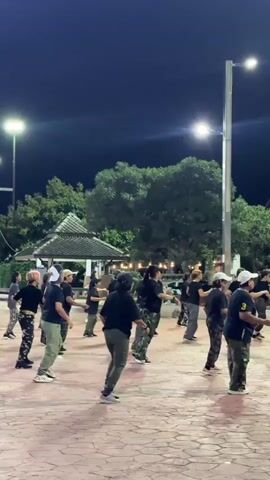 This media file captures an outdoor group exercise or dance class at night. The primary subjects are a group of approximately 20-25 individuals, appearing to be adults, engaged in synchronized movement. They are dressed in casual athletic wear, predominantly black tops and a mix of camouflage and olive green pants.

The setting is an open, paved area with a decorative cobblestone-like pattern. In the background, there are trees, a small, traditional-looking pavilion with a multi-tiered roof, and buildings that suggest an urban or semi-urban environment. Streetlights are visible, illuminating the scene, and a tall lamppost is a prominent feature. The overall impression is that of a public space being utilized for recreational activity after dark.

The activity appears to be a form of aerobic exercise or dance, with participants moving in unison, their bodies angled as if following instructions or a routine. Some individuals are facing forward, while others are turned to the side or back, indicating a dynamic, choreographed movement. The lighting suggests it is nighttime, and the clear visibility implies fair weather. There is no visible text in the media file. The atmosphere seems energetic and focused, with no clear indication of specific emotions beyond the engagement in the activity. The location context provided places this scene in Surat Thani City Municipality, Thailand.