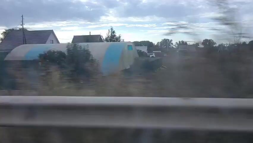 The video captures a blurry, outdoor scene from a moving vehicle, likely a car. The view is through a window with visible water droplets or smudges. In the foreground, a metal guardrail is prominent. Beyond the guardrail, a low, green fence runs horizontally across the frame. Behind the fence, a street or unpaved road is visible, bordered by dry, yellowish grass. Further back, several residential buildings with pitched roofs are scattered amidst trees and vegetation. The sky is overcast with dramatic, cloudy formations. The overall impression is of a rural or semi-urban landscape.