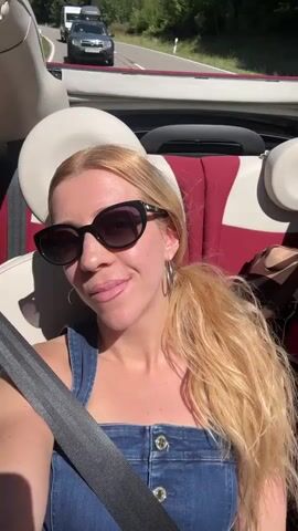 A blonde woman with sunglasses and her hair in a ponytail is in the passenger seat of a convertible. She is wearing a denim jumpsuit and a seatbelt. The top of the car is down, and the interior is red and white. In the background, there are other cars on a road and trees. The woman smiles and makes faces at the camera.