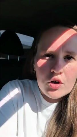 A woman with long brown hair is in the driver's seat of a car. She is wearing a white t-shirt. The car is parked outside. The woman is looking at the camera and talking. In the background, there is a fence, trees, and a house. The sun is shining.