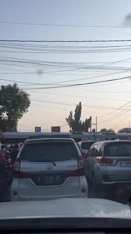 The video shows a first-person perspective of being in a traffic jam. The camera is inside a vehicle, looking forward. In front are several other vehicles, including a white Toyota Avanza with license plate E 1130 CF, and a silver SUV. There are also many motorcycles to the left. In the background, there are trees, utility poles with many wires, and some buildings. The sky is visible above the wires, suggesting it is daytime, possibly late afternoon due to the soft light. The vehicles are stationary, indicating a traffic standstill. There are no clear interactions between people visible, other than drivers being in their cars.