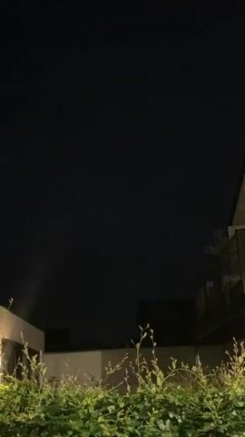 The video shows a night sky with dark clouds. There is a flash of lightning visible in the sky. The foreground is dark, but a beam of light is visible on the left side of the frame. There is also some greenery in the foreground, and part of a building or structure is visible. The video is a short clip, and the lightning strike is brief.