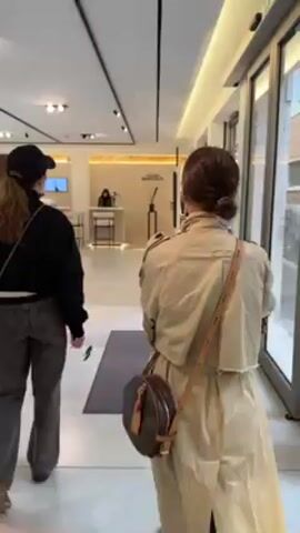 Two women walk into what appears to be an art gallery. The first woman wears a black baseball cap, black jacket, gray jeans, and carries a crossbody bag. The second woman wears a tan trench coat, a brown crossbody bag, and has her hair up in a bun. They walk past a reception desk and a green statue of a woman. The gallery is brightly lit with overhead lights. The floors are light-colored.