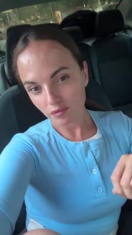 A woman is sitting in the passenger seat of a car, wearing a light blue long-sleeved shirt. She is looking at the camera and moving her head. The interior of the car is visible, including the back seats and headrests. The lighting suggests it is daytime, possibly with some shade from trees visible outside the car windows.