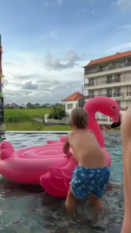 A young child in blue swim trunks tries to get on a pink inflatable flamingo in a pool. The child tries to crawl up onto the flamingo and eventually sits on its back. The pool appears to be an infinity pool with a view of rice paddies and a multi-story building. It is daytime and the sky is blue with scattered clouds.