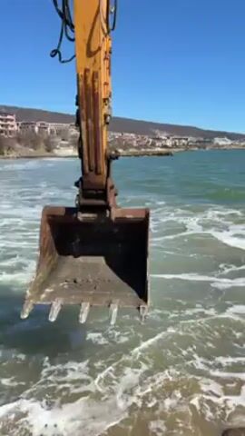The video shows an excavator on a beach, with its bucket submerged in the ocean. The water is turquoise and foamy. A town sits on a hill in the distance under a clear blue sky. The excavator bucket lifts out of the water.