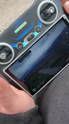 A person operates a drone controller with a built-in screen. The screen displays a live video feed from the drone's camera, showing a mountainous landscape. An error message indicating an "SD card malfunction" is displayed on the screen. The controller has two joysticks, several buttons, and LED indicators. The person's hands are visible holding the controller, and they appear to be manipulating the left joystick. The controller is gray and black with blue accents. The background is a gray surface.