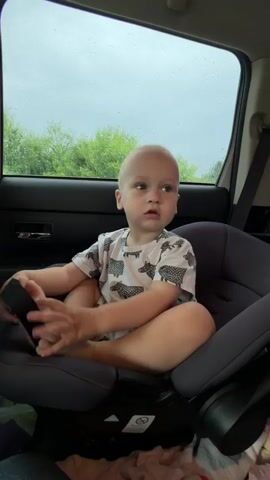 A young child with a bald head is seated in a car seat facing forward. The child is wearing a white t-shirt with a black and white pattern. The child's legs are crossed, and their arms are resting on their lap. The child's hands are spread open. The child is looking to the right with a neutral expression. In the background, there is a window with trees and foliage visible. The window appears to be wet, suggesting it might be raining. The interior of the car is dark.
