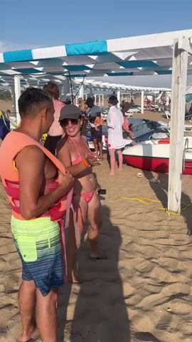 The video shows a group of people on a sandy beach with the ocean in the background. Some people are wearing life vests, suggesting they are about to engage in a water activity like a banana boat ride, which is visible in the distance. People are standing around, talking, and getting ready. The sky is blue with some clouds. There is a white wooden structure providing shade on the beach.