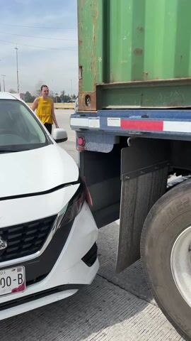 The scene depicts the aftermath of a car accident. A white Nissan, with its front bumper significantly damaged, is positioned directly behind the rear of a large green cargo container mounted on a blue truck trailer. A young woman in a yellow top and dark pants stands behind the car, near a middle-aged man.  A law enforcement officer, in uniform with a vest, is also present and appears to be gesturing with his right hand while holding something small in his left.  The surroundings suggest an outdoor location, possibly a road or highway, with clear skies and power lines visible in the background.
