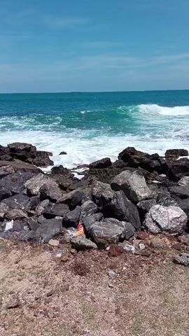 The video shows a rocky coastline under a clear blue sky. The ocean is visible with waves crashing against the rocks. The rocks in the foreground are varied in size and color, with some appearing to be dark grey and others lighter grey with white speckles. There is some litter visible among the rocks, including a plastic wrapper and some small white objects. In the distance, a small island or ship can be seen on the horizon. The camera is positioned at a low angle, looking up towards the ocean and sky.