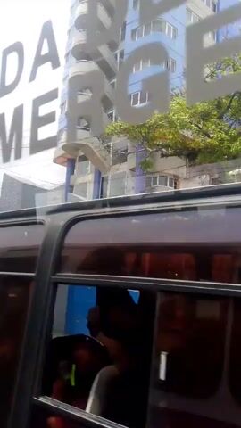 The video is a low-quality, shaky shot from inside a moving vehicle, likely a bus. The focus is on the scenery passing by outside the window.

The initial shot shows the reflection of the letters "PEN DE EMERG" on the bus window, with a tall, light blue and white building visible in the background. The camera then pans to show a building with a blue and red archway and the seal of "Gobernacion Bolivariana de la Guaira." The shot then passes a blue wall with the word "Policia" written on it, and several parked motorcycles are visible behind the wall.

The camera then continues moving, passing a small blue building with a red stripe, a sign for "DAMASCO," and a sign for "ARMATODO." The video ends with a view of a set of stairs leading up to a wall with "CORREDOR WIFI" painted on it. In the background, a hillside and some buildings can be seen.

The overall impression is a quick, mobile view of a street scene in what appears to be a Latin American town. The shaky camera and low video quality make it difficult to discern fine details.