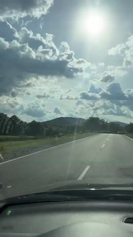 The video shows a view from inside a car driving on a paved road. The sky is partially cloudy, with the sun shining brightly. In the distance, there are rolling hills covered in trees. The main focus of the video is a large flock of sheep crossing the road. The car slows down as it approaches the sheep, allowing them to cross safely. The sheep are mostly gray or white in color. The road has a solid white line in the center. There is a "no entry" road sign visible on the right side of the road.