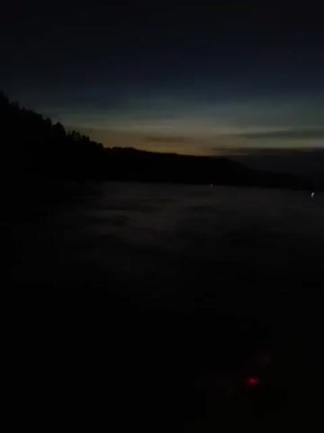 The video shows a body of water at dusk or dawn, with a dark silhouette of land on the left horizon. The sky is a deep blue, transitioning to lighter shades near the horizon, suggesting the sun is either rising or setting. The water appears to be choppy with subtle ripples. A portion of a boat's railing is visible in the foreground of the latter half of the video, along with colored lights reflecting on the water below. A tall, thin pole extends vertically from the boat. The scene is very dark, with limited visibility.