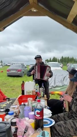 A medium shot shows a man standing outdoors under a wooden canopy, playing an accordion and singing. He is wearing a black cap, a black jacket, and a dark shirt. In front of him is a wooden table laden with various items including bottles, plastic containers, mugs, and food. To his right, two individuals are seated at the table, one wearing camouflage clothing and holding a thermos, the other partially visible. In the background, a grassy field is visible with several cars parked under a cloudy sky. A white tent is also visible in the background.