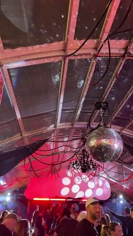 The video shows a crowded indoor event space, likely a party or concert, with a large disco ball hanging from the ceiling. The ceiling is made of a reflective material with a grid-like structure, and is illuminated by red lights and white circular shapes that appear to be part of a decorative display. Red cables are strung across the ceiling. Below the disco ball, a black chandelier is also visible. In the foreground, a crowd of people are gathered, many facing away from the camera, suggesting they are watching a performance or engaged in conversation. Some individuals are holding drinks. The overall atmosphere appears to be lively and festive.