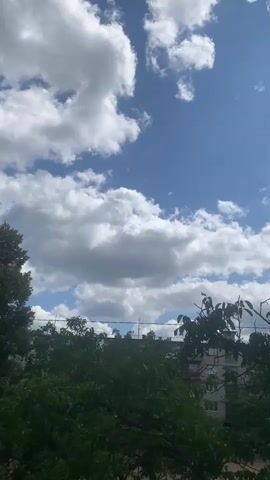 A vertical video shot from a high vantage point looks up at a bright blue sky filled with fluffy white clouds. Below the sky, the tops of trees with lush green leaves are visible, some obscuring the view of buildings in the background. The buildings appear to be residential, with several stories and visible windows. Power lines crisscross the scene above the trees. The lighting suggests it is daytime, likely morning or afternoon, with the sun illuminating the clouds and foliage. The overall impression is of an urban or suburban environment on a clear day with some cloud cover.