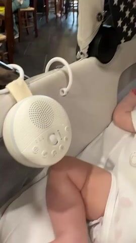 A white portable sound machine with multiple buttons and lights hangs from a stroller. A baby's leg and arm are visible in the foreground, lying on a white surface. The background is blurred, showing a dimly lit indoor setting with chairs and tables. The sound machine is playing, as indicated by the green light.