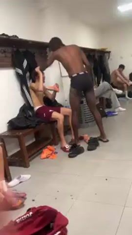 The video shows a locker room with several men present. Two men are the central focus. One man is sitting on a wooden bench, shirtless, wearing maroon shorts, and appearing to be focused on his feet. Another man stands near him, shirtless and wearing black underwear, his attention also on the floor near his feet. The surrounding floor is littered with shoes, sandals, and scattered debris. A maroon gym bag is visible in the foreground. In the background, there are other men in various states of undress, sitting on benches, and a laundry basket. The locker room itself is sparsely furnished, with lockers lining the wall and a mirror present. Overall, the scene conveys the aftermath of some physical activity, likely a sporting event or practice session.