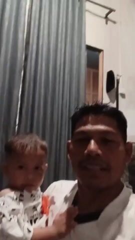 A man with short dark hair and a mustache holds a young child with short brown hair. The man is wearing a white shirt with a black collar. The child is wearing a white shirt with a black and white pattern of what appear to be cartoon animals. They are indoors in front of a grey curtain. A fan is visible behind the man on the right side of the frame.
