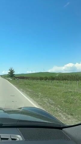 The video shows a car driving along a narrow road on a sunny day with a clear blue sky. In the distance, there are green fields with windmills. There is a white guardrail along the side of the road. The road has a white line dividing the two lanes.