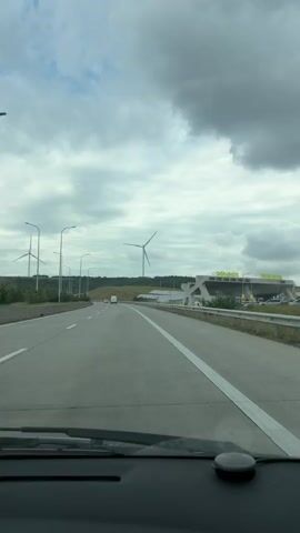 The video is shot from the perspective of a driver on a highway. The highway is divided into multiple lanes with white markings. On the right side of the highway, there is a toll plaza with a modern, angular structure. Beyond the toll plaza, there are several buildings and structures. In the distance, on the left side of the highway, there are numerous wind turbines with their blades slowly rotating. The sky is overcast with dramatic clouds. A few vehicles are visible on the highway, and one truck appears to be leaving a plume of white smoke or steam. The dashboard of the car is partially visible in the foreground.