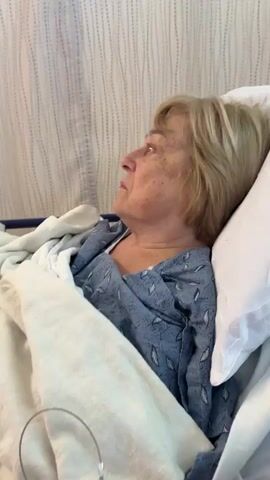 The video shows a close-up of an older woman lying in a bed. She is wearing a blue and white patterned hospital gown and is covered with a white blanket. Her hair is blonde and shoulder-length. She is looking to her left at the beginning of the video, then turns to look directly at the camera with a distressed expression. Her eyes are blue and appear to be slightly red, suggesting she may have been crying. The background consists of a light-colored, textured wall, possibly a hospital room. The lighting is soft and appears to be coming from above.