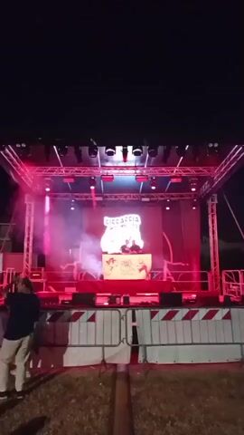 A nighttime outdoor concert stage is illuminated with bright red lights and smoke. A DJ is visible on stage behind a console, with a logo displayed on a screen behind them. A large banner with text and a logo hangs from a metal structure to the right of the stage. Fencing and barriers are set up in front of the stage.