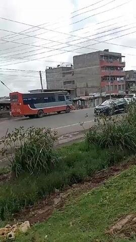 The video shows a roadside scene in what appears to be an urban or semi-urban setting. A black SUV is driving on the road, followed by an orange and gray bus. On the side of the road, there is green vegetation. Buildings are visible in the background, and power lines are overhead. The overall impression is of a typical street scene with traffic.