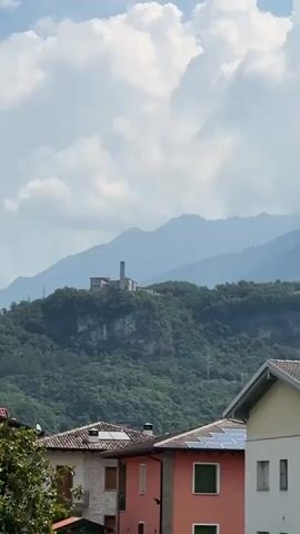 This video displays a picturesque mountainous landscape with a building on a hilltop. The foreground features residential buildings with tiled roofs, some with solar panels, and a streetlamp. The middle ground is dominated by a lush green, tree-covered hillside leading up to the prominent building. In the background, layers of misty blue mountains recede into the distance under a sky filled with scattered white clouds. The overall scene evokes a sense of tranquility and the beauty of a rural Italian setting, possibly in Cividate Camuno, Italy, given the location context. The lighting suggests it is daytime, likely late morning or early afternoon, with the sun filtering through the clouds. There are no visible people or any specific activity depicted; the focus is on the static scenery. The building on the hilltop appears to be a structure with a tower, possibly a church or historical monument, standing out against the natural backdrop.