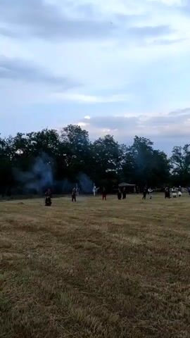 A wide shot depicts an open, dry grassy field under an overcast sky with scattered clouds. In the mid-ground, a line of approximately 20-30 individuals, some dressed in dark historical military-style uniforms, are positioned in front of a treeline. Multiple plumes of white smoke erupt from their locations, coinciding with the sounds of several loud gunshots, indicating the firing of prop firearms or muskets. The camera slowly pans horizontally from left to right, revealing more participants and onlookers gathered along the treeline, and eventually a building in the distant background. The ground is covered in short, dry, cut grass.