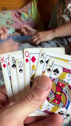 A first-person perspective video captures a hand holding playing cards, with the cards being King of Diamonds, 8 of Spades, 10 of Spades, Queen of Diamonds, King of Spades, and Jack of Spades. In the background, two other people are visible, one on the left and one on the right. The person on the left is handling cards and appears to be dealing or playing. The person on the right is also interacting with cards. The setting seems to be indoors, possibly on a bed, with a wooden wall and bedding visible. The overall activity appears to be a card game.