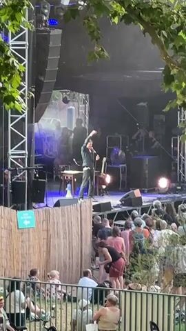 A man in a dark outfit and a cap stands on a raised platform at an outdoor concert, speaking into a microphone and gesturing with his hand. He has a guitar propped up beside him. The stage behind him is illuminated with blue and white lights, and other figures are visible on the stage, some appearing to be musicians. A bamboo fence runs along the front of the platform, separating it from an audience seated on the ground and standing behind the fence. The audience members are diverse in age and are looking towards the stage. Trees and foliage hang over the top of the frame, suggesting the concert is in a natural or park-like setting.