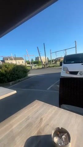 The video opens with a view from under an awning, showing two wooden tables and a wicker chair in the immediate foreground, suggesting a patio setting. Beyond, a street leads to a railroad crossing marked by a red and white barrier. A person walks briefly past the crossing at the beginning of the clip. Buildings line the background under a clear, bright blue sky.

At 0:01, the camera pans right to reveal a white Renault van parked in a lot. The pan continues, and at 0:02, a white Audi Q5 SUV, displaying the French license plate "GM-626-HR," becomes visible, parked beside the van. The sun is low in the frame, creating significant lens flare and a bright glare.

Across the street, several buildings are visible, including one prominently marked with a green cross and the word "PHARMACIE" (Pharmacy). The video concludes at 0:05, showing both the white Renault van and the white Audi Q5 parked on the sunny street. The weather is clear and bright throughout the recording, indicating either morning or late afternoon.