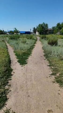A dirt path splits into two forks in a grassy field under a clear blue sky. The camera moves forward along the right fork of the path. In the distance, there are some buildings, including one with a blue roof. Trees and vegetation line the sides of the path. The video appears to be taken on a sunny day.